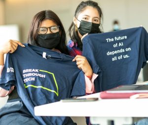two women hold up break through tech T-shirts that say "the future of AI depends on all of us"